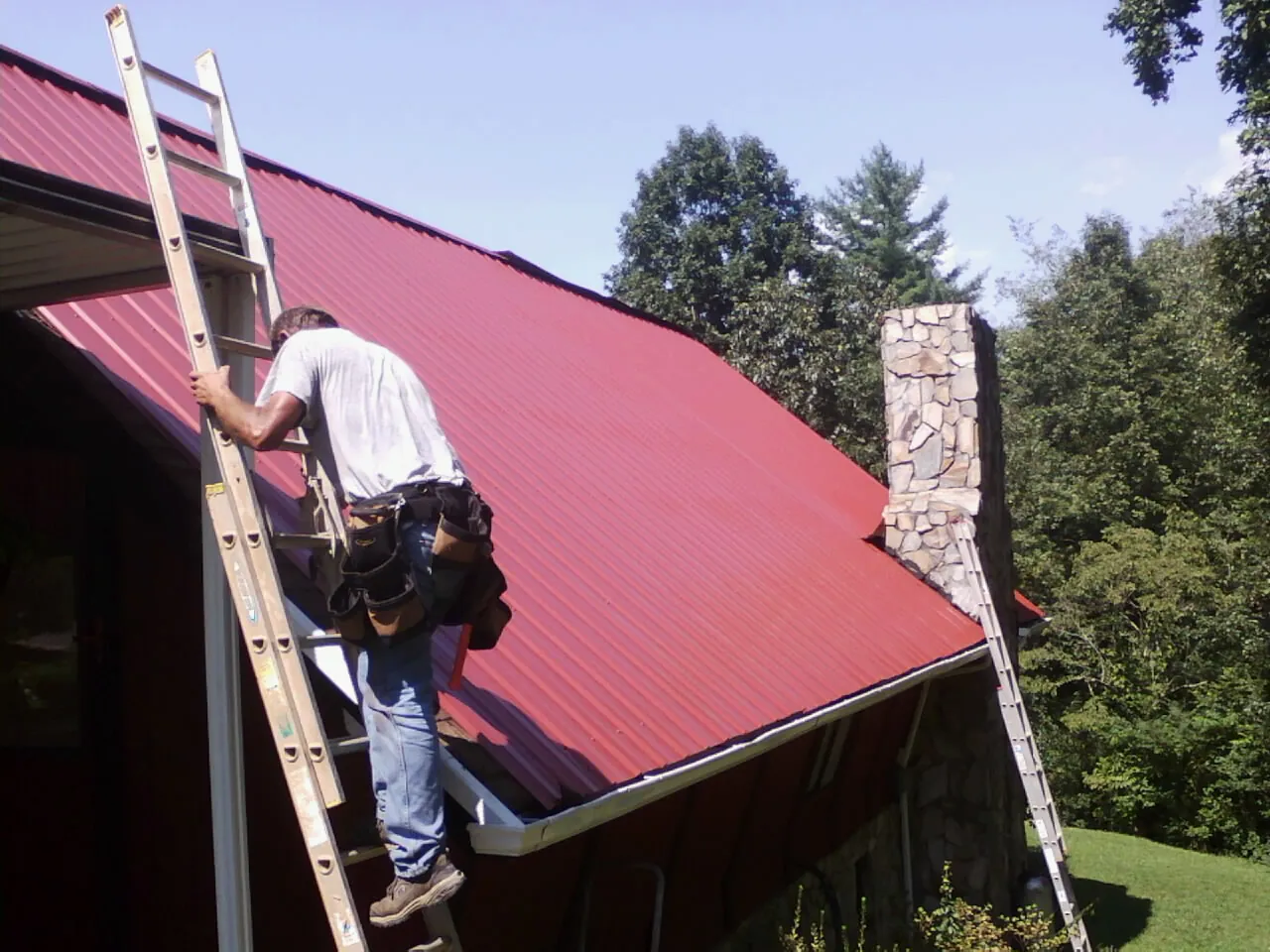 Roofer on a ladder beside a red metal roof and stone chimney — Soffit & Fascia Repair by Roofing Brothers in New Lenox